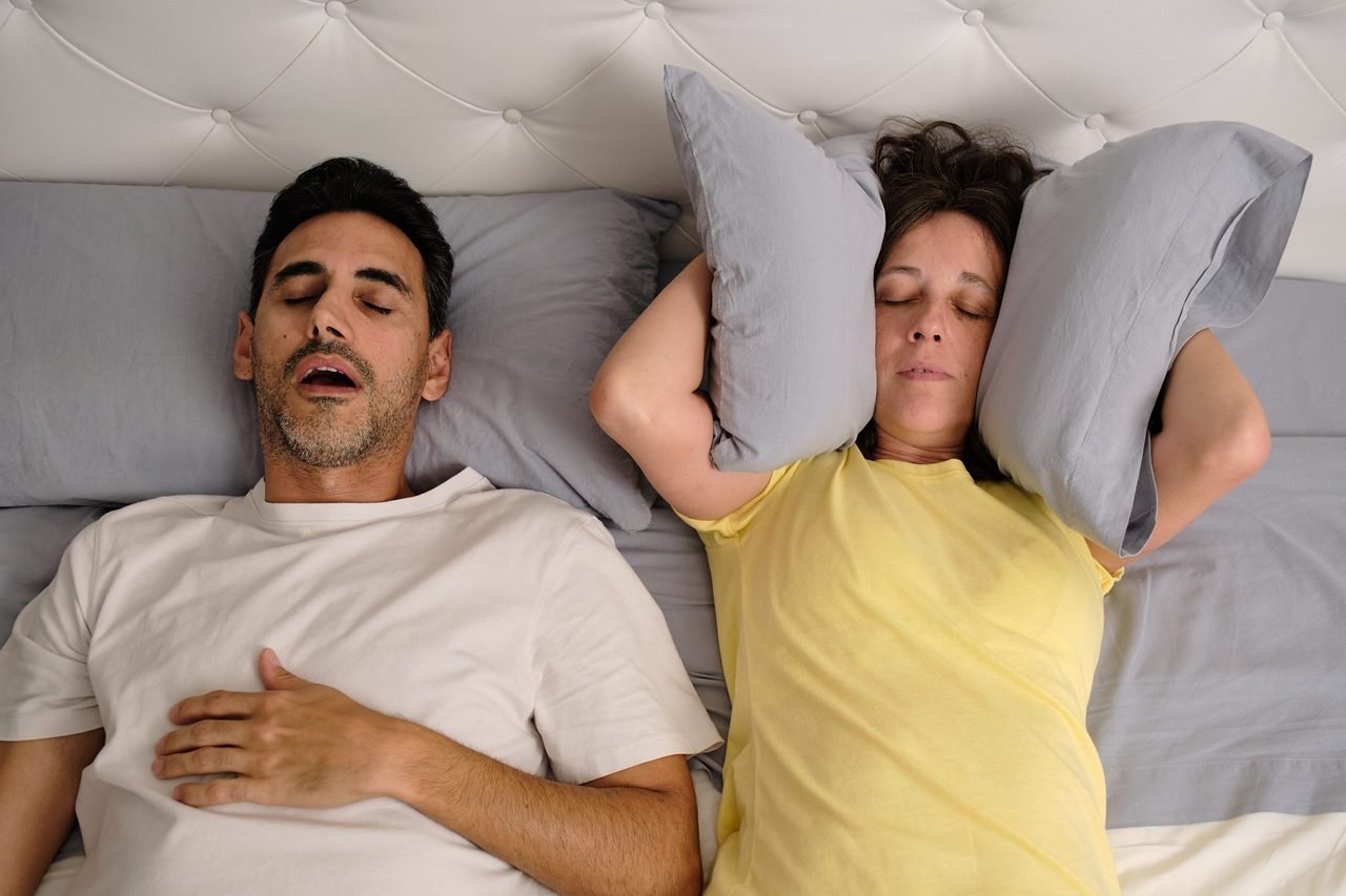 Man snoring on his back in bed while his partner presses pillows over her ears