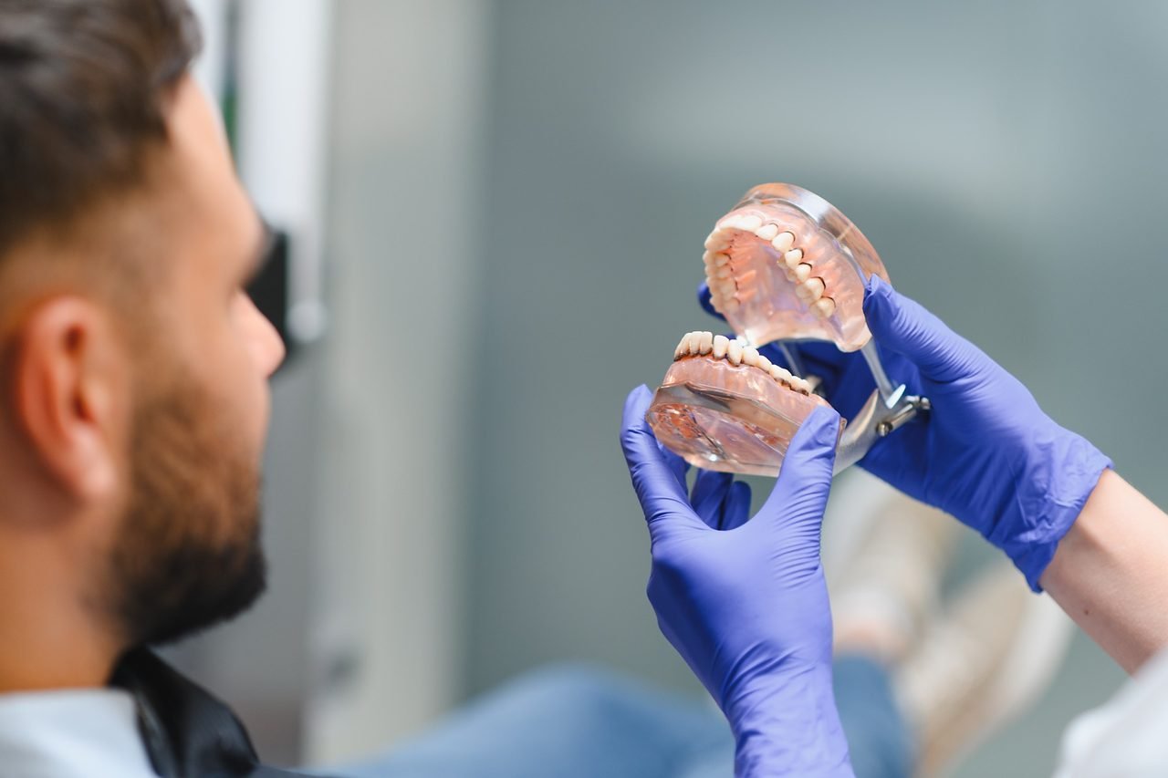 Dentist in blue gloves showing a jaw model to a patient in the dental chair